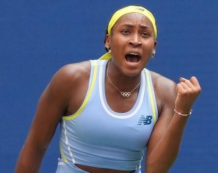 Coco Gauff celebrująca zwycięstwo na korcie tenisowym, z uniesionymi rękami i szerokim uśmiechem, na tle wiwatującego tłumu i tablicy wyników.