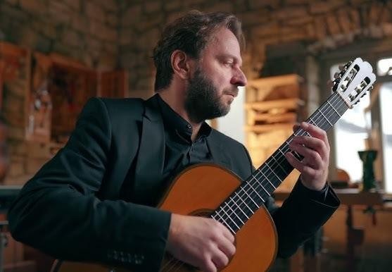 Zbliżenie na dłonie Marcina Dylli grającego na gitarze klasycznej, podkreślające precyzję palców na gryfie i delikatne ruchy strun, w tle rozmyta atmosfera sali koncertowej.