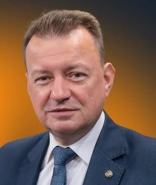 Mariusz Błaszczak przemawiający z mównicy lub w trakcie spotkania politycznego, ukazujący jego rolę jako aktywnego polityka.