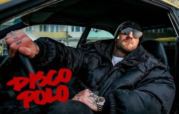 Kizo (Patryk Woziński) w zbliżeniu, patrzący wprost na widza, w swobodnym otoczeniu, z naturalnym oświetleniem.
