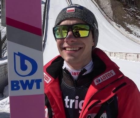 Jakub Wolny w kombinezonie skoczka narciarskiego, z nartami, na tle ośnieżonej skoczni.